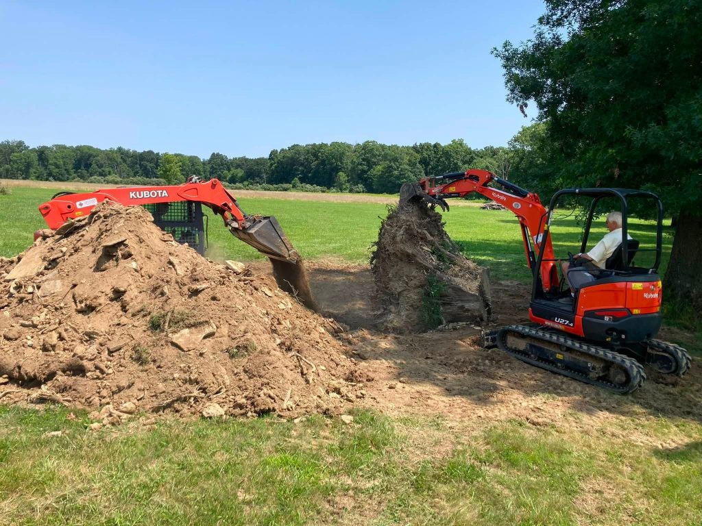 Mini excavator clearing dirt beside a large removed tree stump in an open field